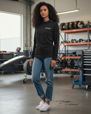 Woman wearing a black Crown Speed Shop Ultra hooded long sleeve shirt with blue jeans in an automotive garage.